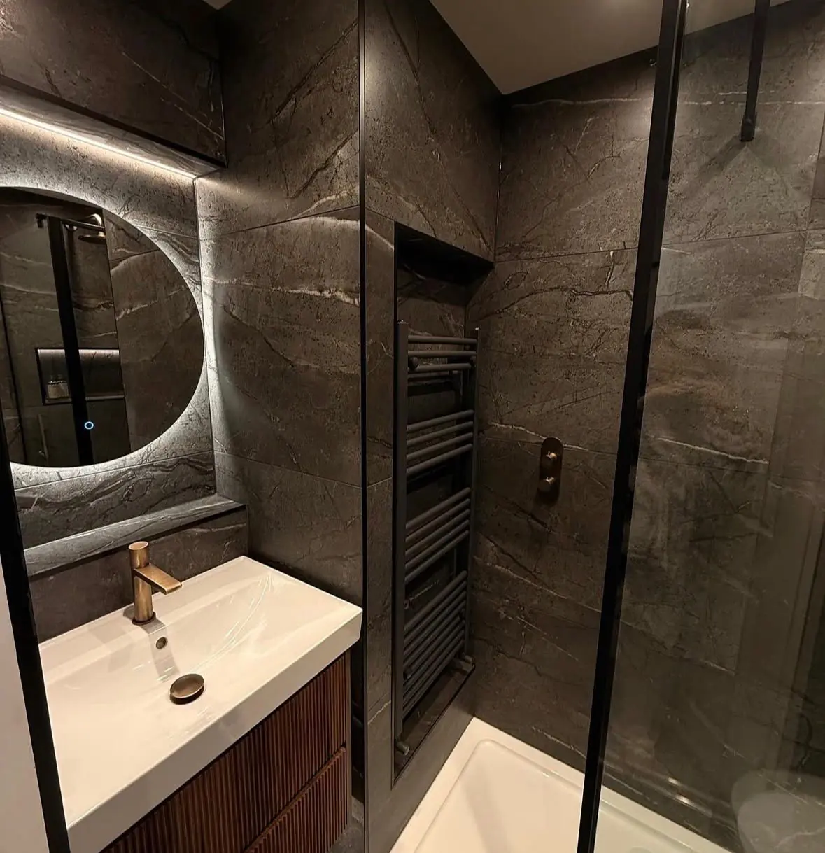 Compact shower room in dark veined stone tiles with a round backlit mirror and a brass tap